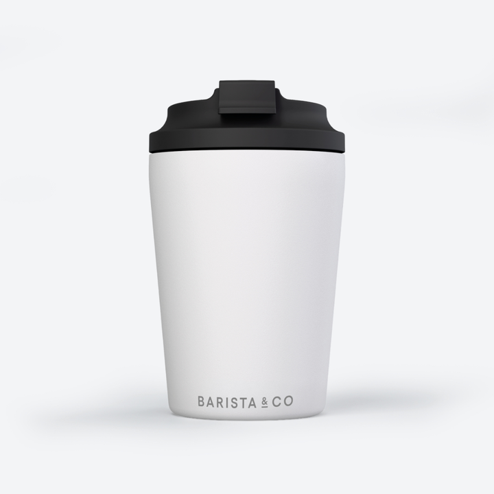 A white travel tumbler with a black flip-top lid, featuring the 'BARISTA & CO' logo printed in black near the bottom, set against a clean white background.