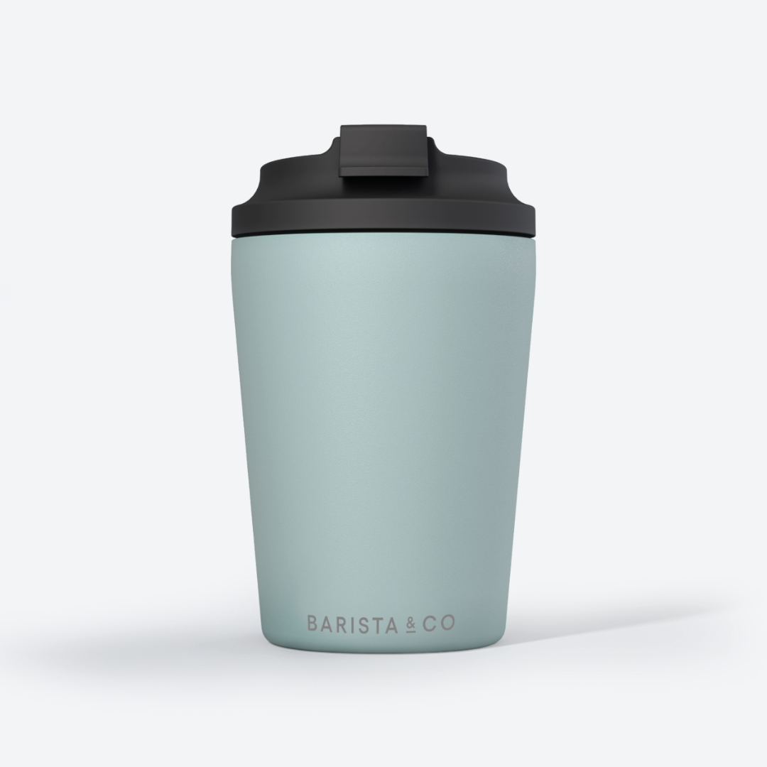 A light sage green insulated travel tumbler with a black lid, featuring the 'BARISTA & CO' logo printed in gold near the bottom, set against a clean white background.