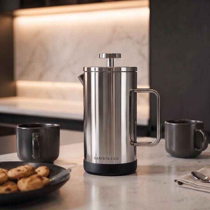 A stainless steel French press coffee maker with a black base and handle, set on a marble countertop in a modern kitchen setting with dark mugs and cookies nearby.