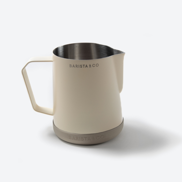 A Barista & Co. stainless steel milk pitcher with a cream-colored handle and body, a brushed silver base, and black printed branding on the side, set against a clean white background.
