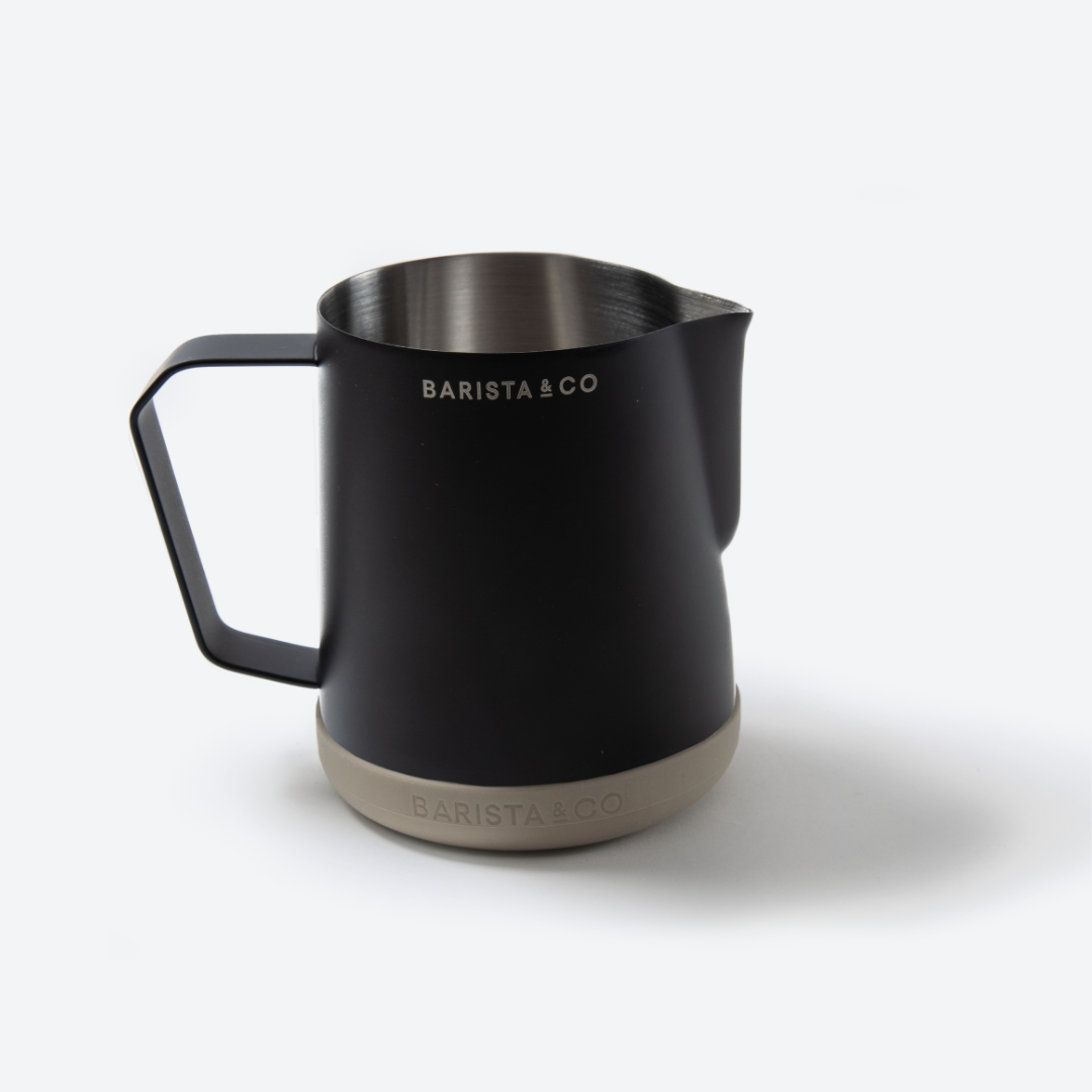 A matte black stainless steel milk pitcher with a black handle, featuring a brushed silver interior, white 'BARISTA & CO' branding on the upper body, and a beige textured base.